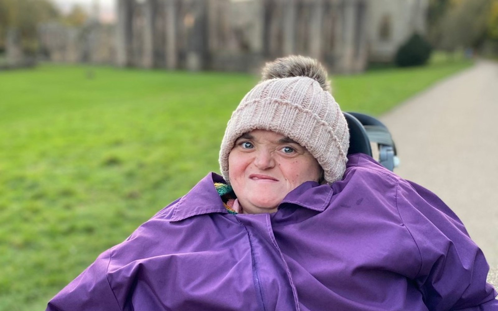 Daisy smiling with a winter coat and hat on, with green grass behind her 