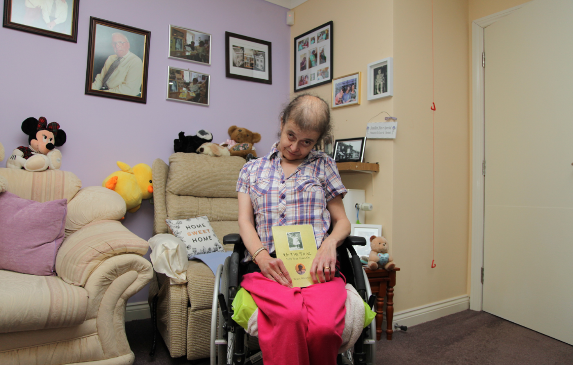 Linda smiling from her wheelchair holding a yellow book named "up the trail, fifty four years on" written by Linda herself